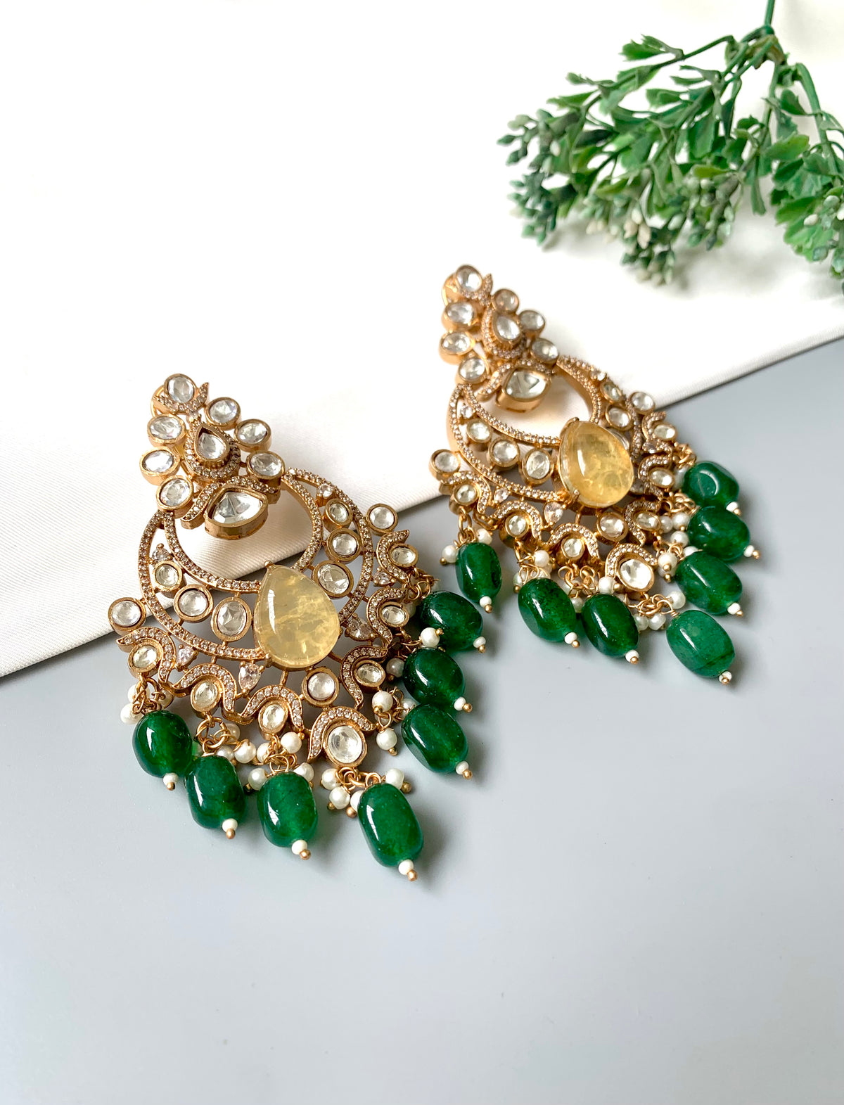 A pair of ornate gold-toned chandelier earrings decorated with clear stones, a central yellow stone, and dangling green beads, displayed on a light gray surface with a white card and green leaves in the background.