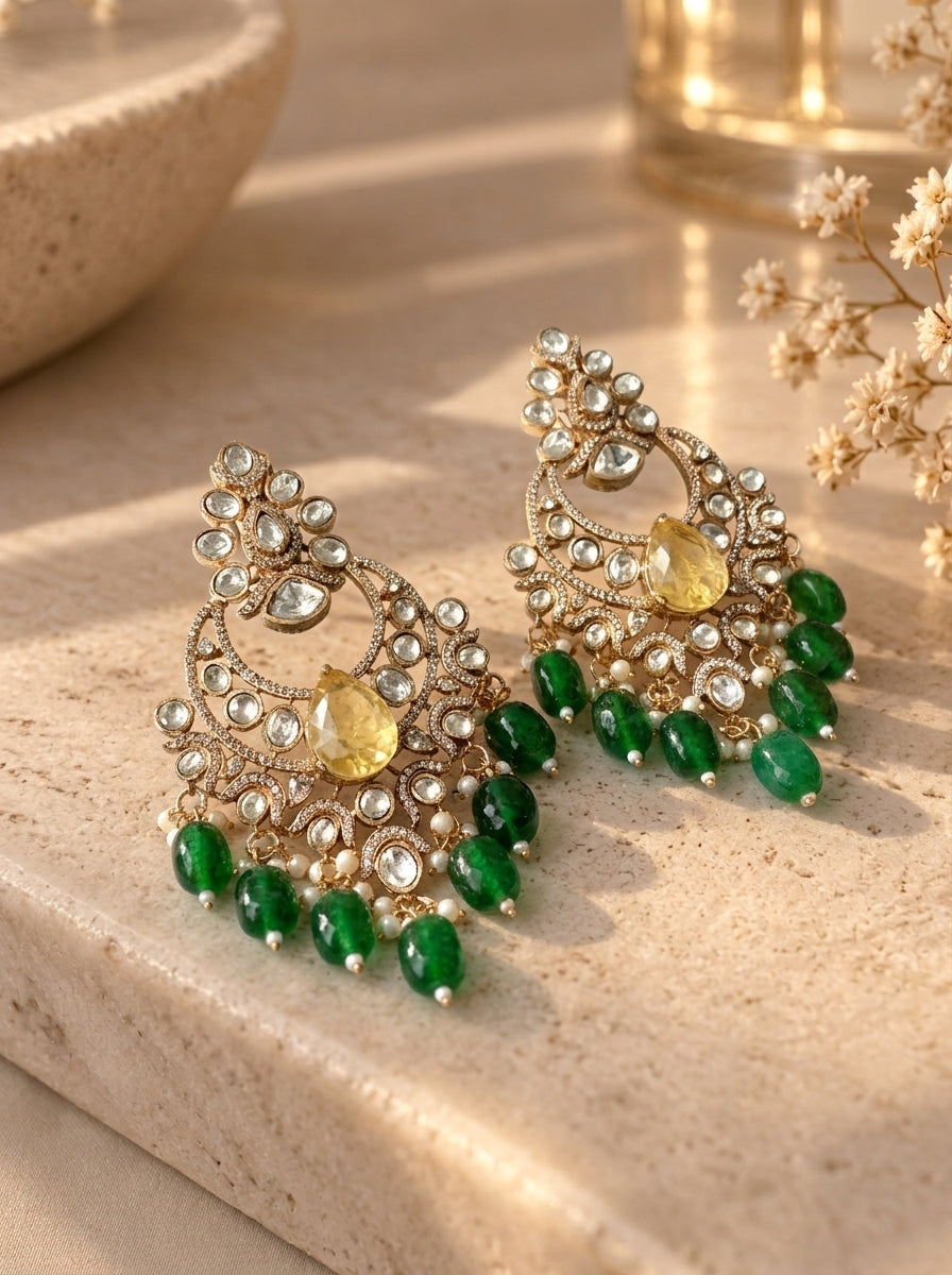 A pair of ornate gold-toned chandelier earrings with clear stones, yellow center stones, and dangling green bead drops, displayed on a beige stone surface with warm natural light.