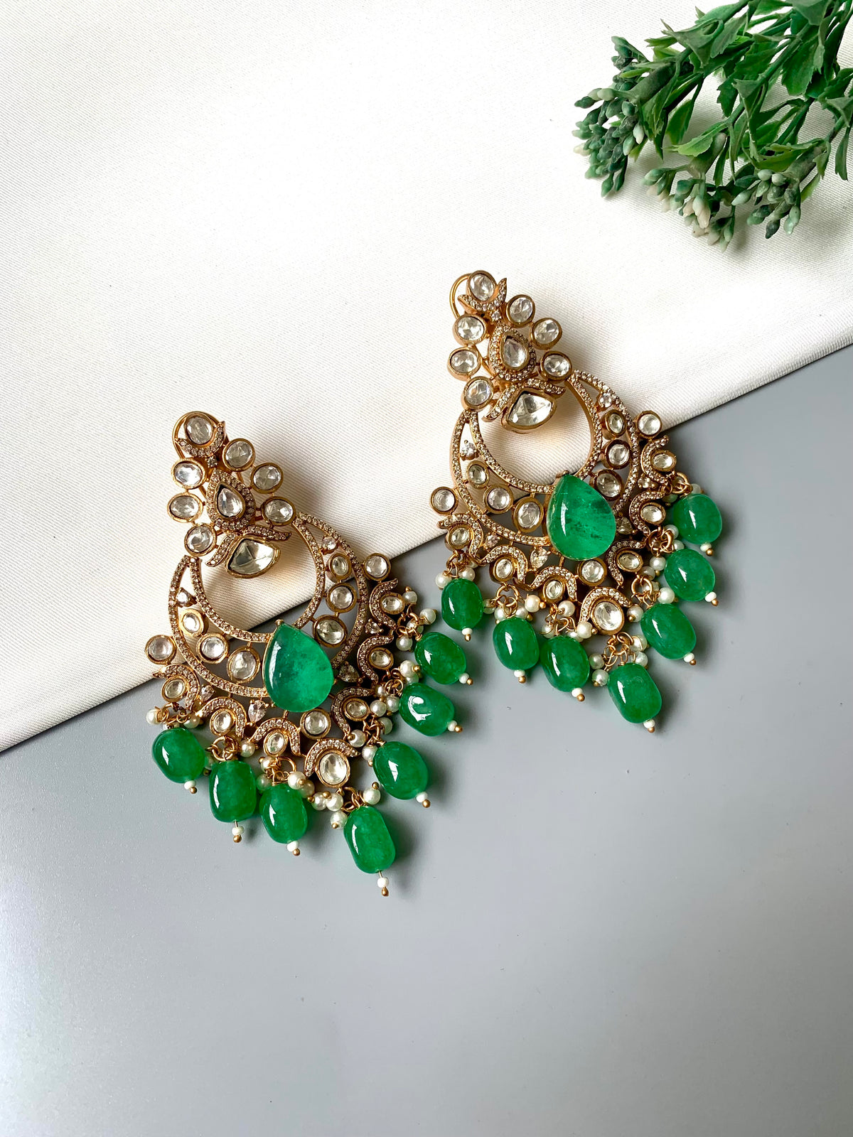 A pair of ornate gold-toned chandelier earrings with green gemstone accents and small clear stones, displayed on a white and gray surface with a small green plant in the corner.