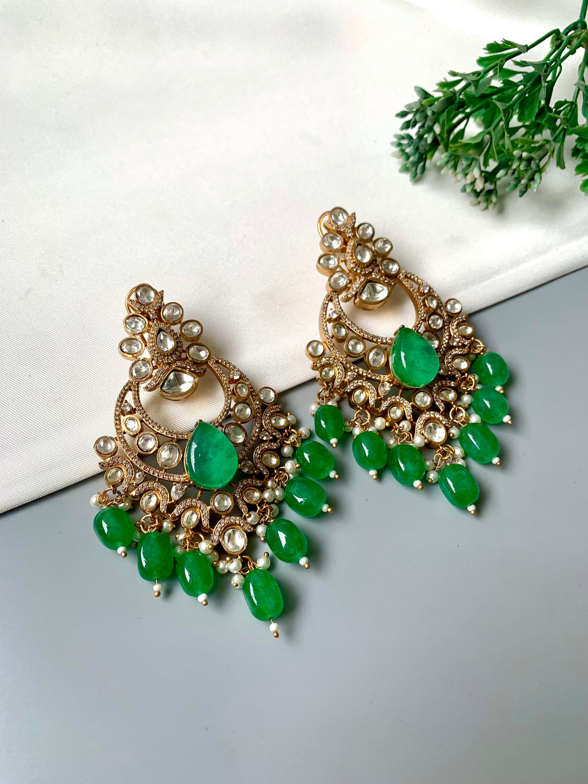 A pair of ornate gold-toned chandelier earrings with green gemstone accents and small pearl-like details, displayed on a light gray surface with a white fabric prop and green leaves in the corner.