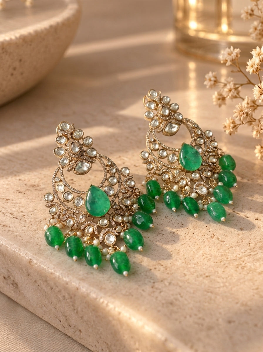 A pair of ornate gold-toned chandelier earrings with green gemstone accents, displayed on a beige stone surface with warm sunlight and dried flowers in the background.