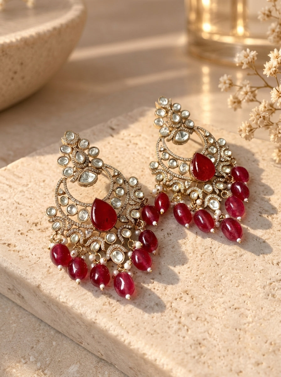 A pair of ornate gold-toned chandelier earrings embellished with clear stones and dangling red gemstones, displayed on a beige stone surface with warm natural light.