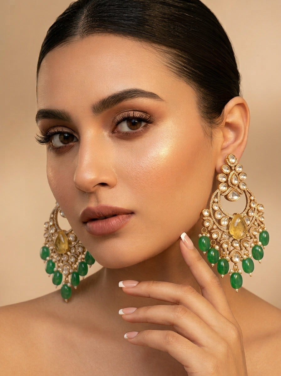A close-up portrait of a woman wearing large ornate gold and green chandelier earrings, with her hand near her face against a warm beige background.