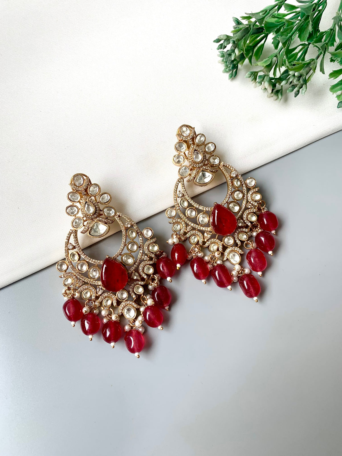 A pair of ornate gold-toned chandelier earrings decorated with clear stones and red bead drops, displayed on a light gray surface with a white card and green leaves in the corner.