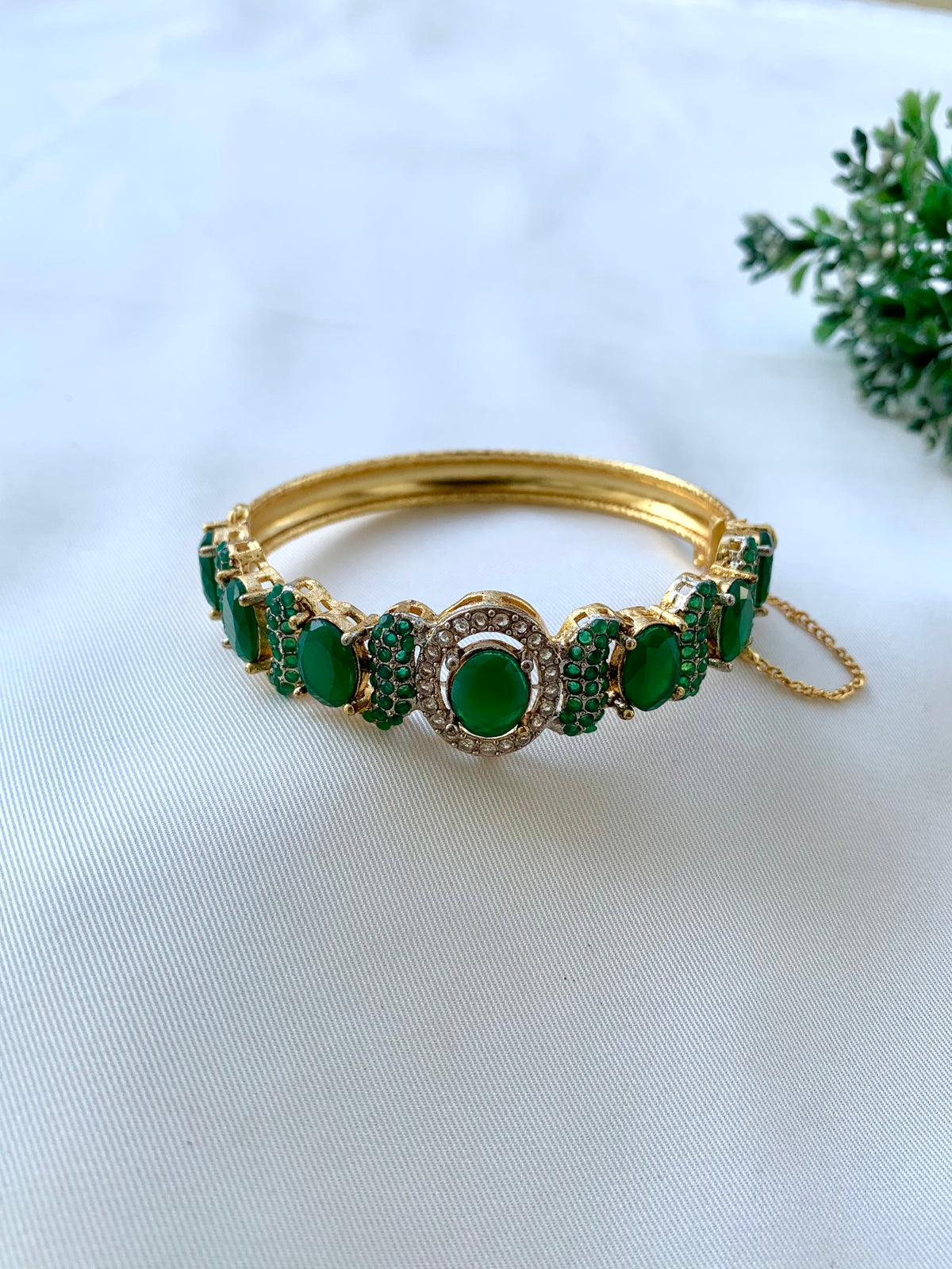 A gold-plated bangle bracelet adorned with multiple green oval cabochon gemstones and small green pavé accents, set against a white textured fabric background with a small green leafy sprig in the corner.