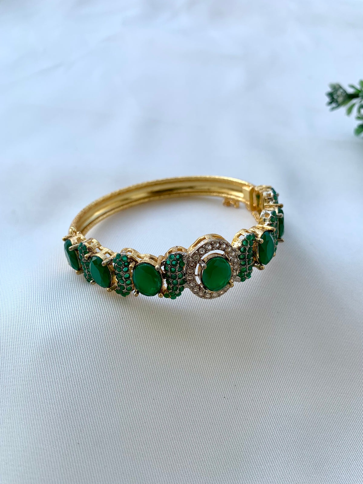 A gold-plated bangle bracelet adorned with green oval and round cabochon gemstones, set against a white fabric background.