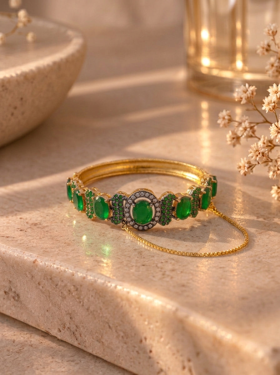 A gold bangle bracelet with green emerald-cut gemstones and a delicate gold chain, displayed on a beige stone surface with warm, soft-focus background elements.
