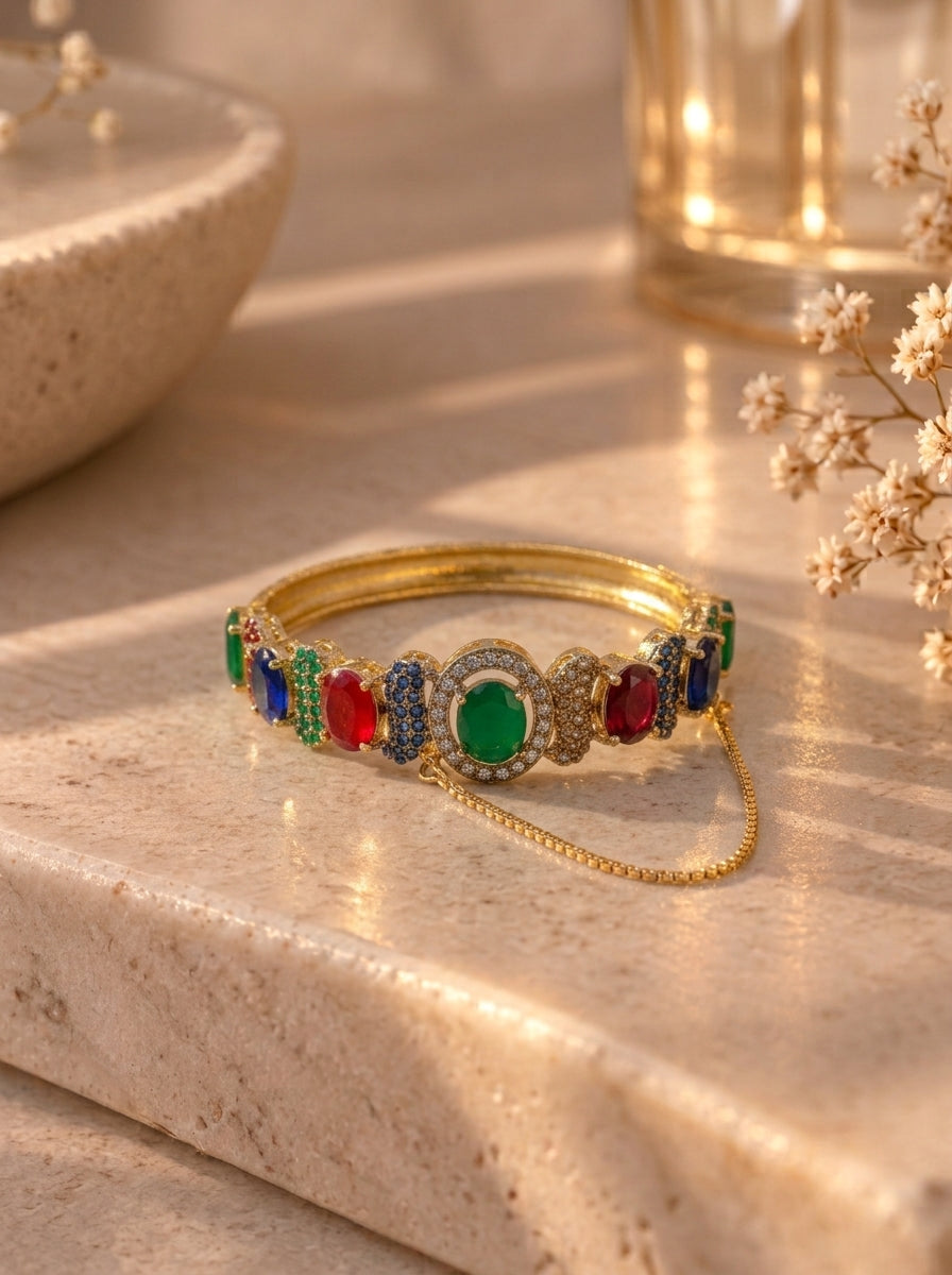 A gold-plated bangle bracelet decorated with colorful gemstones, including green, red, and blue, resting on a beige stone surface with warm sunlight and dried flowers in the background.