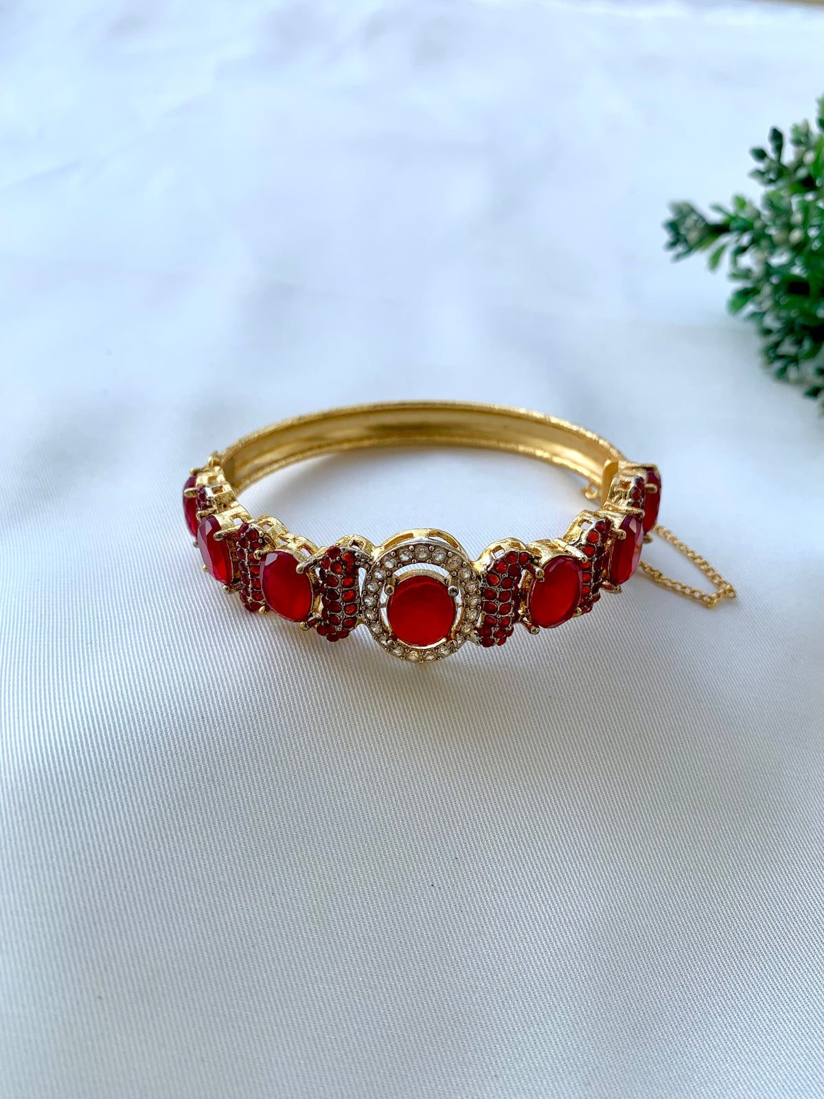 A gold-plated bangle bracelet adorned with red oval and marquise-cut crystals, accented with small clear stones, resting on a white textured fabric background.