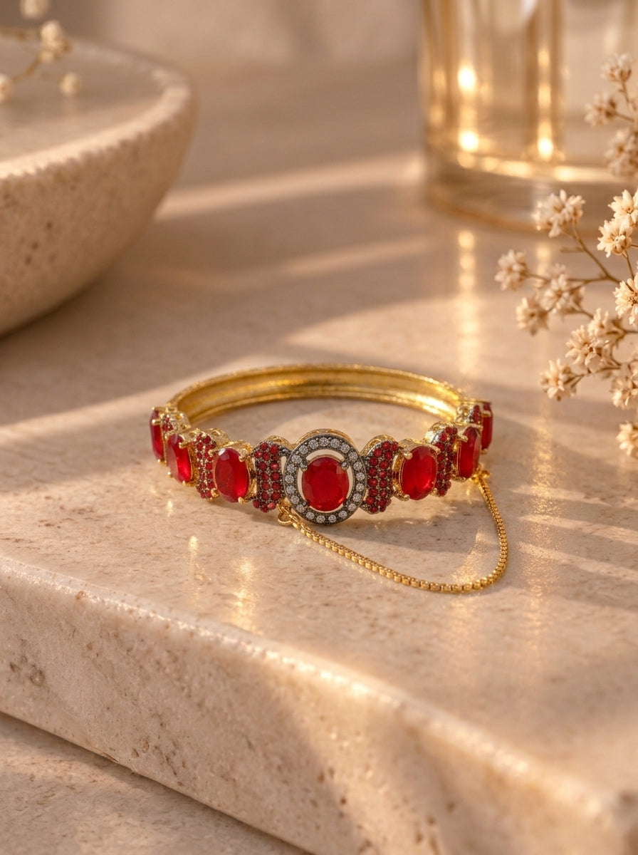 A gold-plated bangle bracelet decorated with oval red gemstones and a central round red stone, accented with small clear stones, resting on a beige stone surface with warm sunlight and dried flowers in the background.