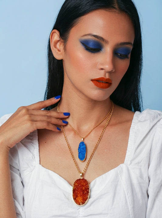 A woman with dark hair, blue eyeshadow, and orange lipstick wears two gold necklaces: one with a rectangular blue stone pendant and one with a larger oval amber-orange stone pendant, set against a light blue background.