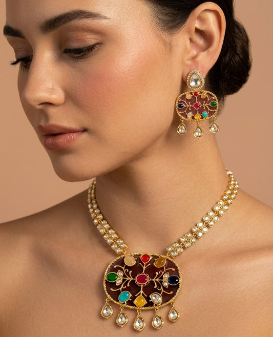 A close-up of a woman wearing an ornate gold-toned jewelry set featuring a statement necklace and matching drop earrings with multicolored stones and pearl-like accents, against a warm beige background.