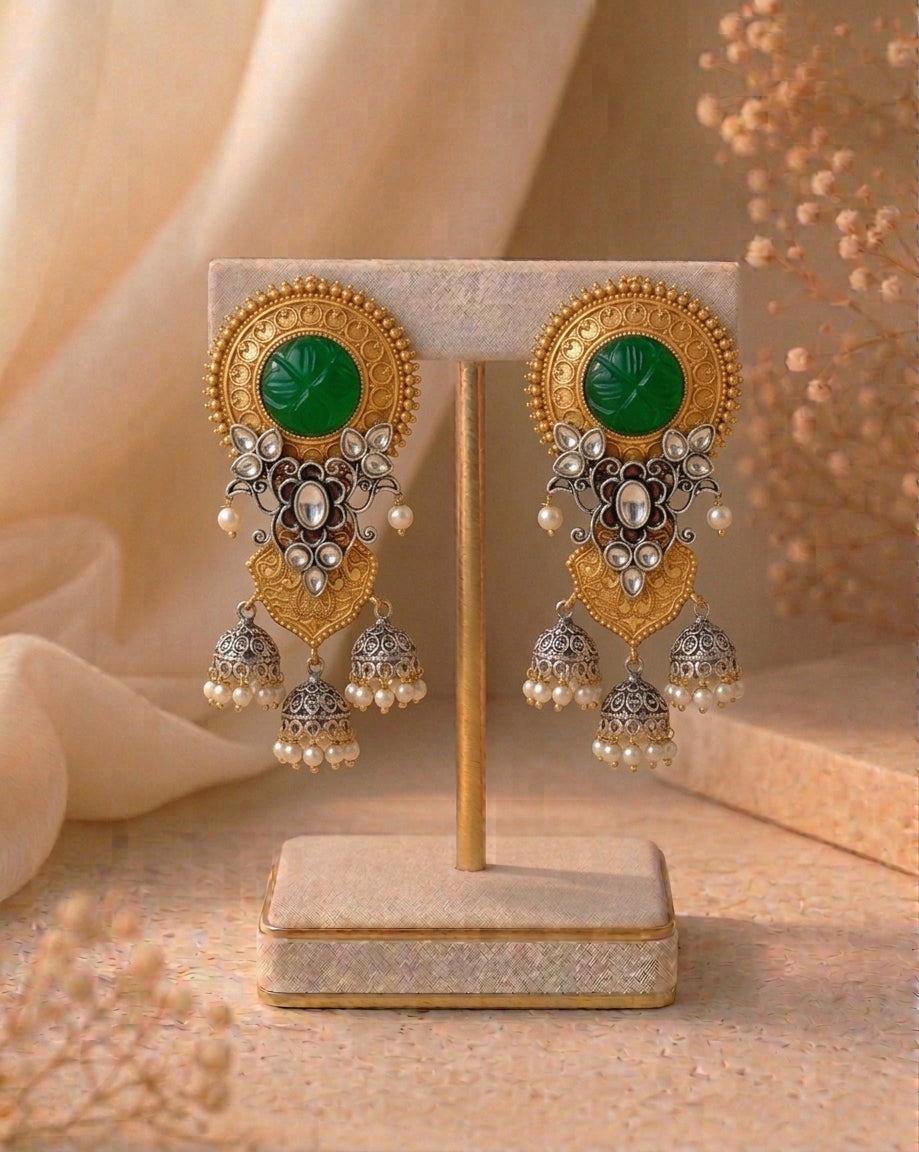 A pair of ornate Indian-style gold and silver dangle earrings with green cabochon stones and white pearl accents, displayed on a beige jewelry stand.