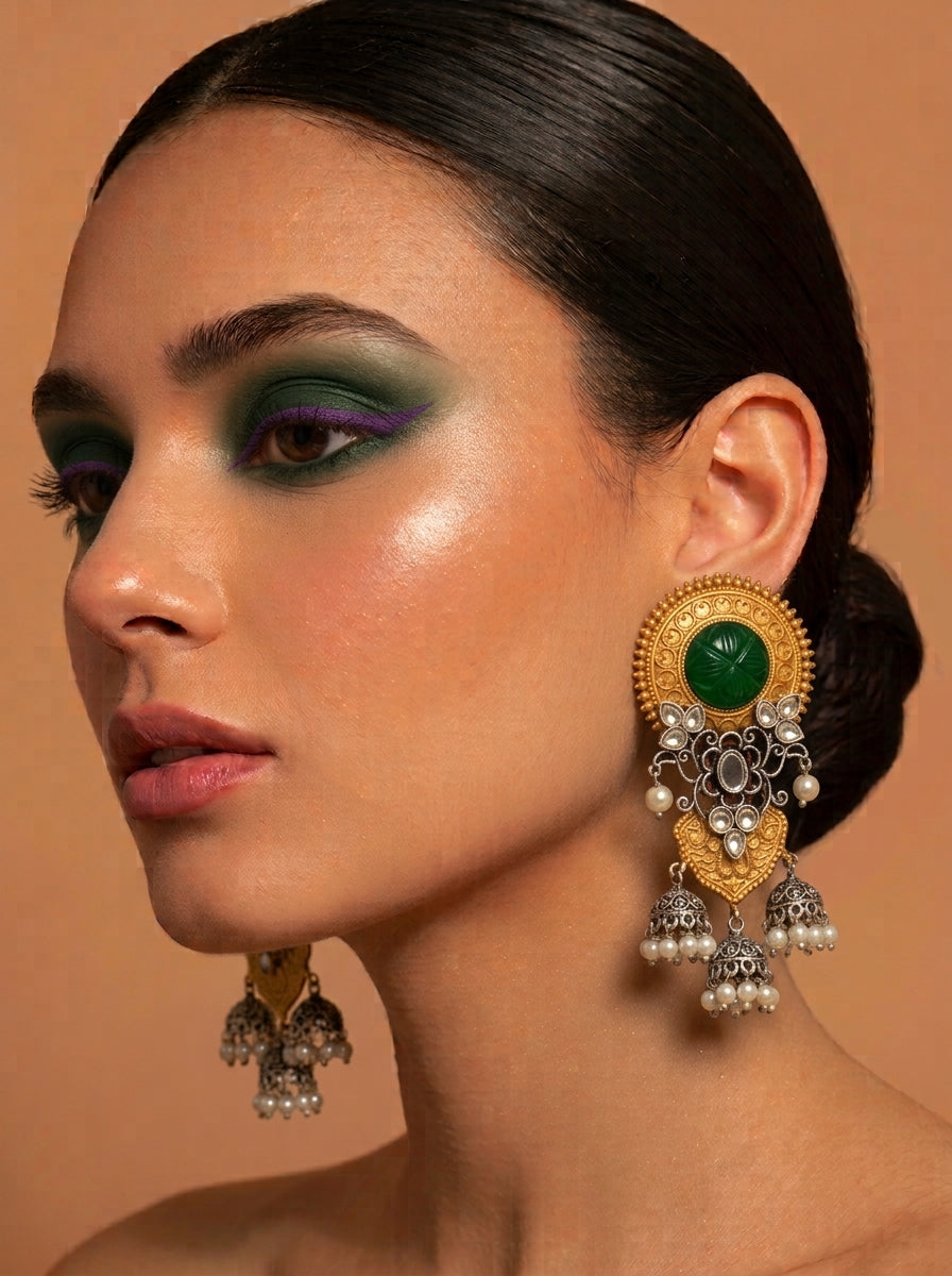 A close-up of a woman wearing an ornate gold and silver jhumka earring with a prominent green stone, set against a warm beige background.