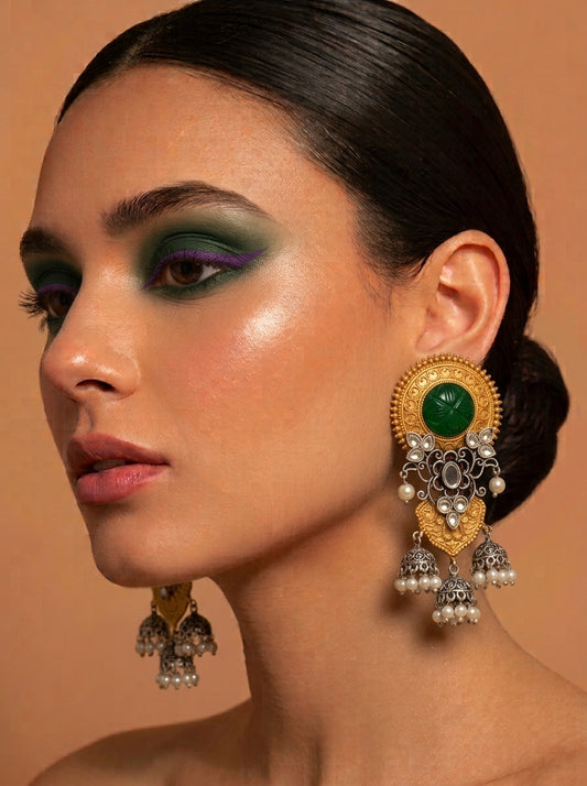 A close-up of a woman wearing an ornate gold and silver jhumka earring with a prominent green stone, set against a warm beige background.