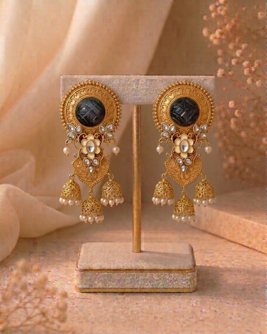 A pair of ornate gold-toned dangle earrings displayed on a beige jewelry stand. Each earring features a round black stone, floral gold detailing, small white pearl-like beads, and dangling bell-shaped jhumka elements.
