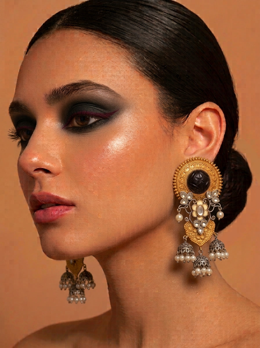 A close-up of a woman with dark hair pulled back, wearing an ornate gold and silver jhumka earring with a black stone and white pearls, against a warm beige background.