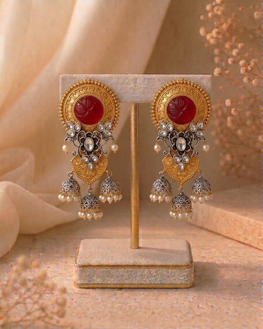 A pair of ornate gold-toned Indian jhumka earrings with red cabochon stones and silver-toned filigree details, displayed on a beige jewelry stand against a warm beige background.