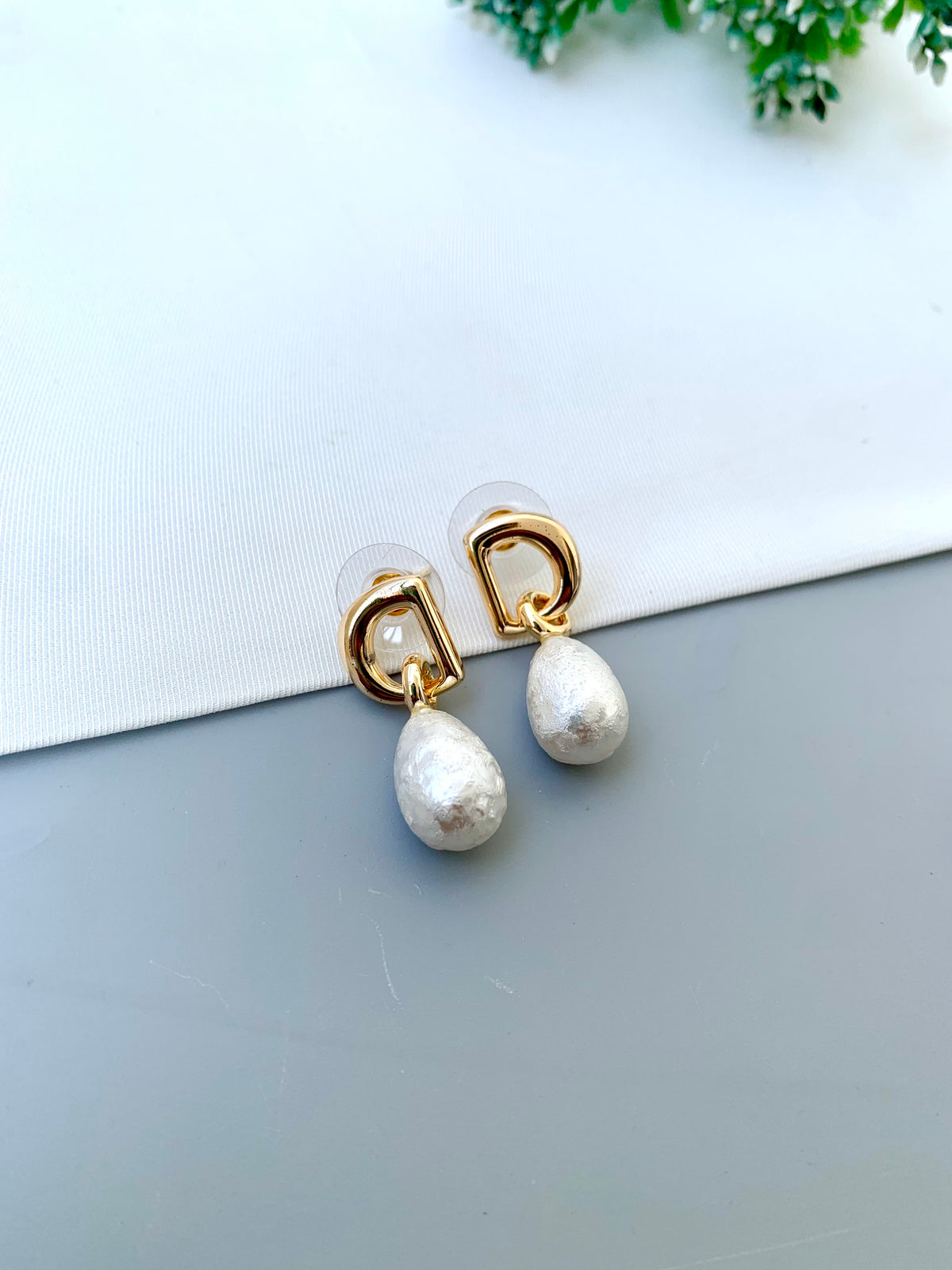 A pair of gold and white pearl drop earrings with a geometric gold frame design, displayed on a split white and gray surface.