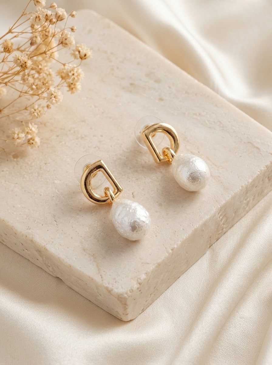 A pair of gold hoop earrings with dangling white pearl drops, displayed on a textured cream stone slab with dried baby's breath flowers and cream satin fabric in the background.
