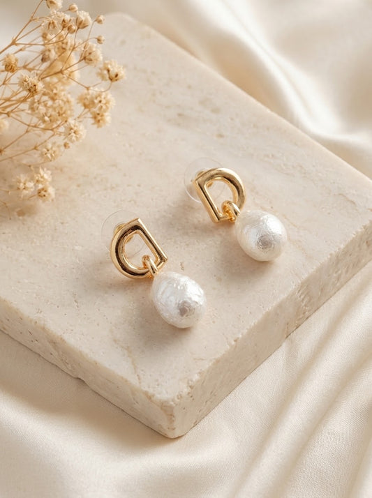 A pair of gold hoop earrings with dangling white pearl drops, displayed on a textured cream stone slab with dried baby's breath flowers and cream satin fabric in the background.
