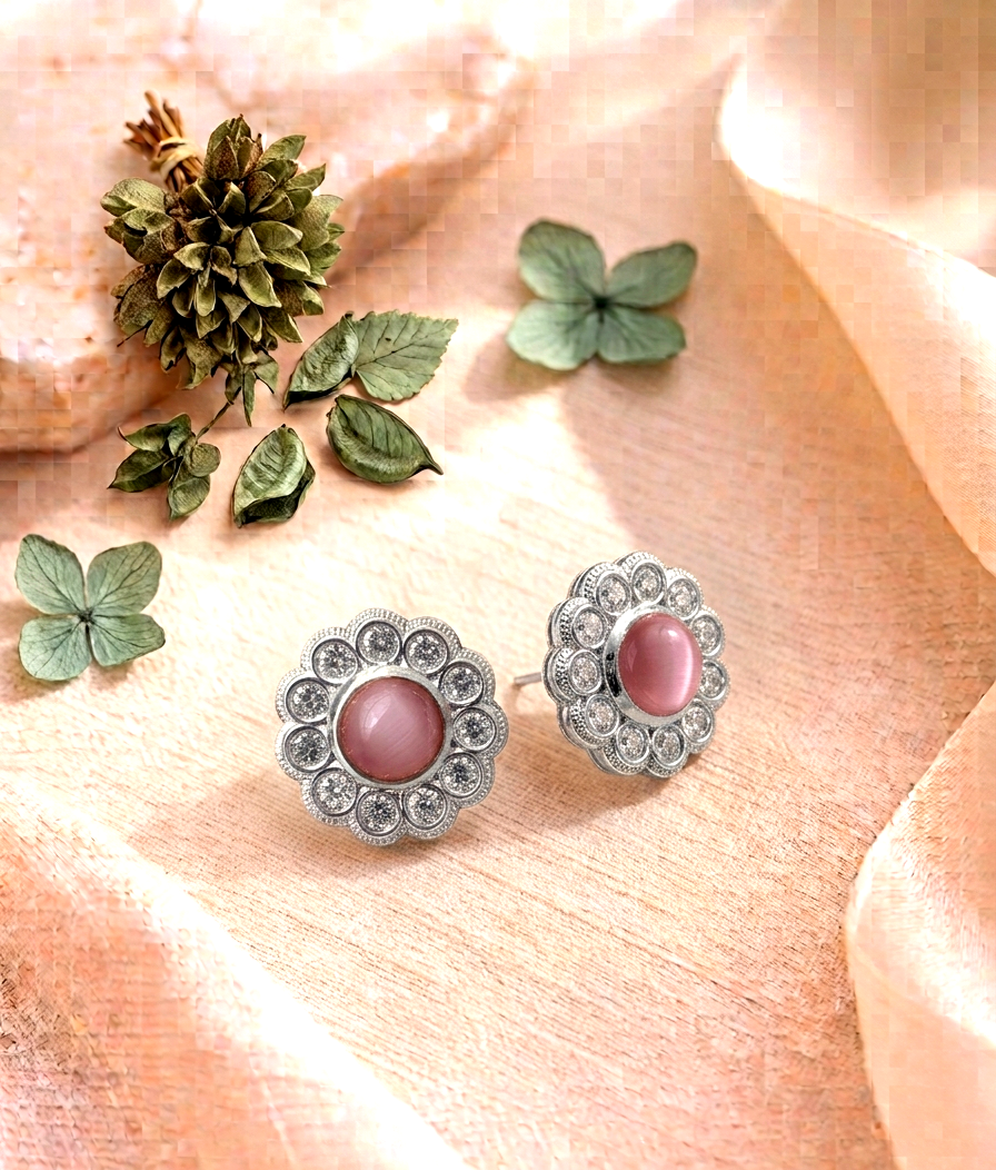 A pair of silver-toned floral stud earrings with oval pink cabochon centers, arranged on peach satin fabric with decorative green leaves and a small pine cone.