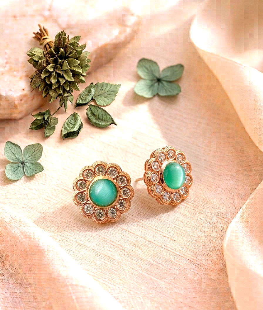 A pair of gold-toned floral stud earrings with oval green cabochon centers and clear rhinestone accents, arranged on soft peach fabric with decorative green leaves and a small pine cone.