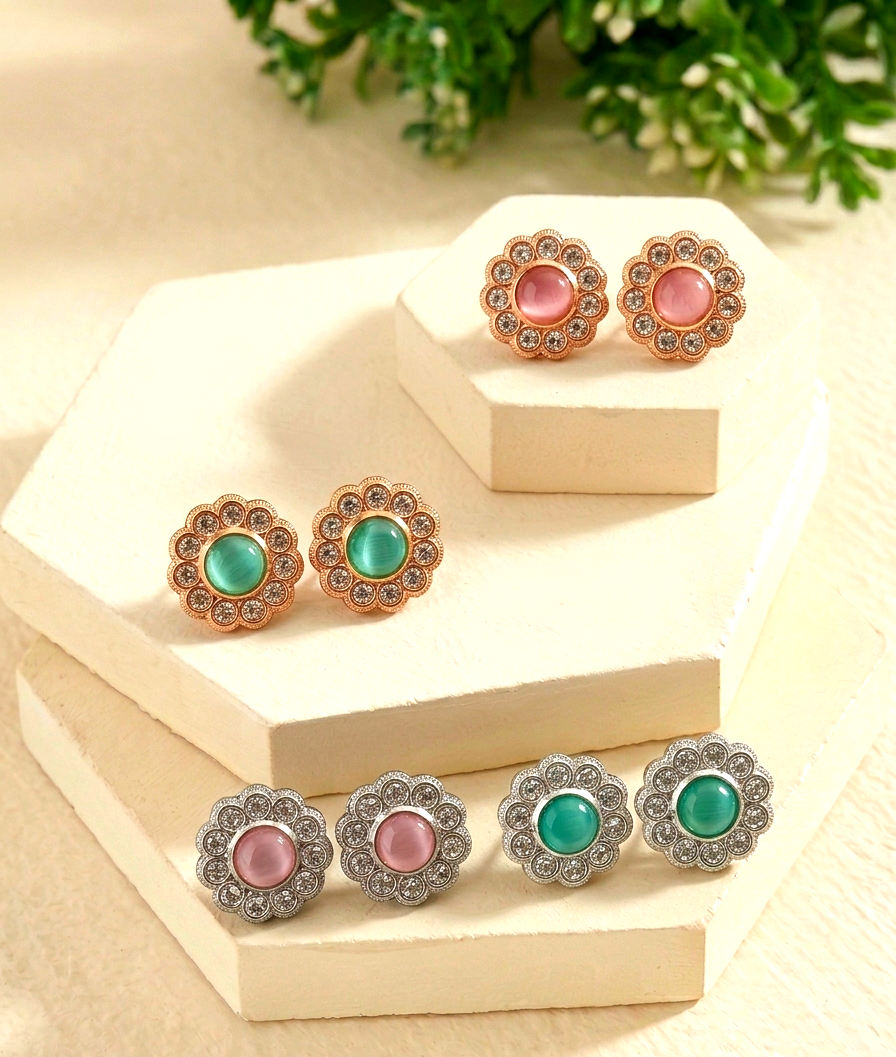 Four pairs of floral stud earrings displayed on cream-colored geometric blocks. The earrings feature round central stones in pink and green, surrounded by small clear rhinestones in rose gold and silver-tone settings.