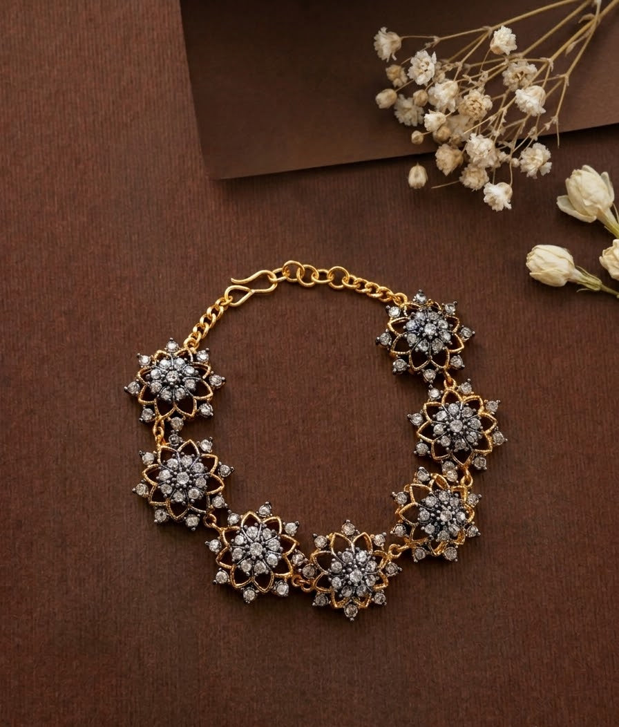 A gold-toned statement necklace featuring five large, intricate floral cluster motifs set with clear stones, displayed on a textured dark brown background with dried white flowers.