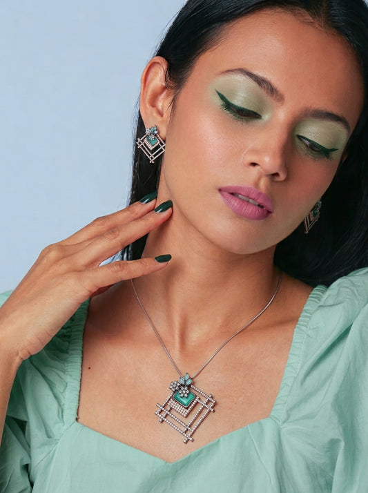 A close-up of a woman wearing a silver geometric pendant necklace with a green stone, matching geometric earrings, and a mint green top against a light blue background.