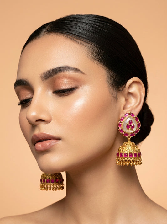 A close-up portrait of a woman wearing ornate gold jhumka earrings with pink gemstone accents, set against a solid warm beige background.