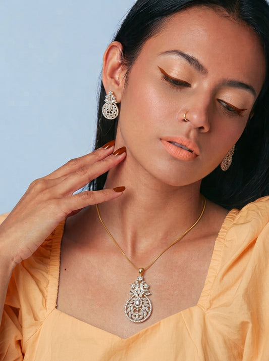 A woman wearing a gold chain necklace with a large silver-toned filigree pendant and matching drop earrings, styled with a peach-colored top against a light blue background.