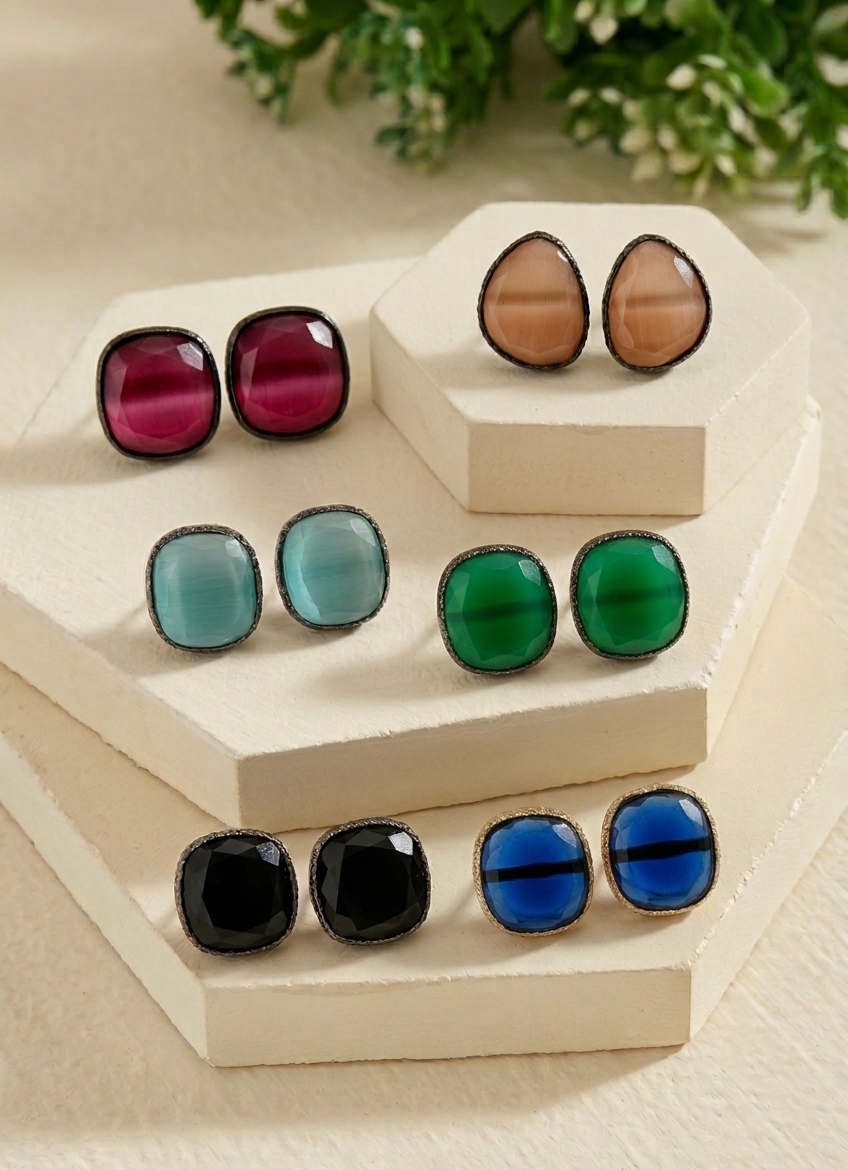 Nine pairs of square stud earrings with smooth, glossy cabochon stones in various colors (pink, blue, green, black, and brown) with dark metal settings, arranged on a beige geometric display stand.