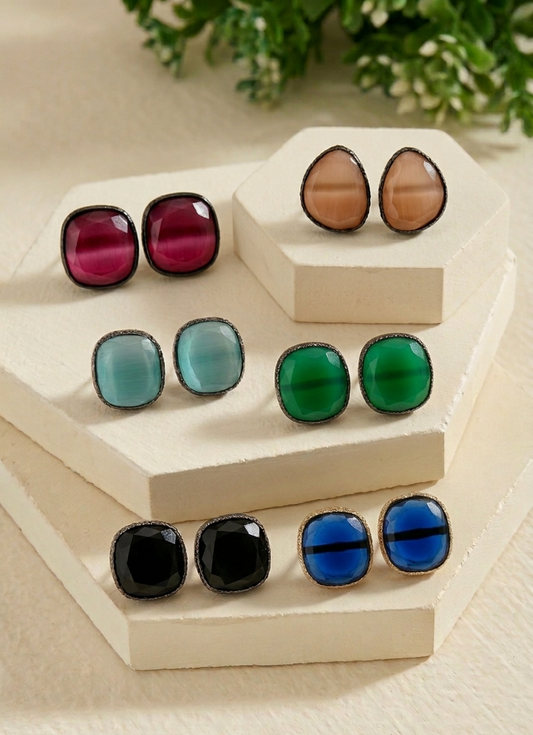 Nine pairs of square stud earrings with smooth, glossy cabochon stones in various colors (pink, blue, green, black, and brown) with dark metal settings, arranged on a beige geometric display stand.