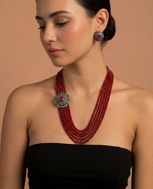 A woman wearing a multi-strand red beaded necklace with a large silver-toned circular pendant featuring a red center stone, paired with matching drop earrings, against a warm beige background.