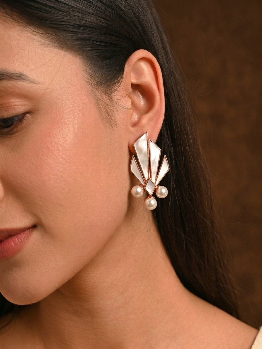 A close-up of a woman's ear wearing a statement earring with layered fan-shaped silver-toned metal and three small pearl-like beads at the bottom, against a warm brown background.