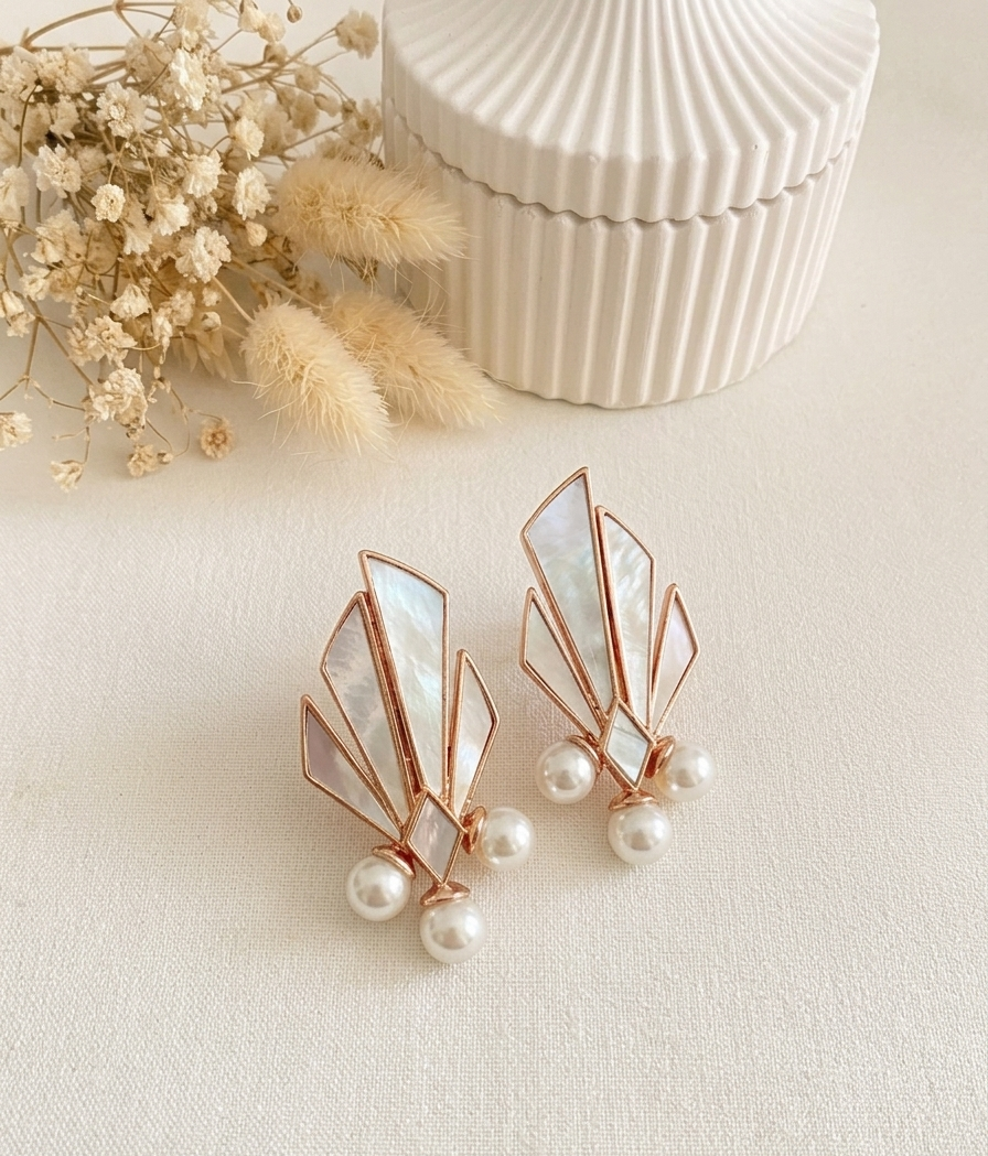 Rose gold geometric stud earrings with iridescent mother-of-pearl inlays and dangling pearl accents, styled on a cream surface with dried flowers and a ribbed cream box in the background.