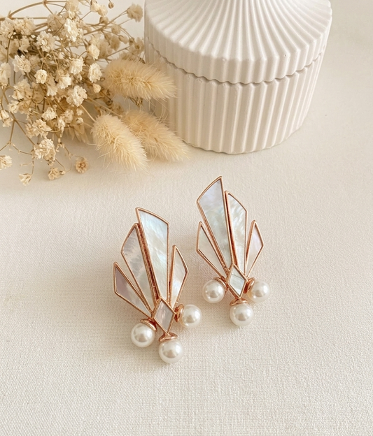 Rose gold geometric stud earrings with iridescent mother-of-pearl inlays and dangling pearl accents, styled on a cream surface with dried flowers and a ribbed cream box in the background.