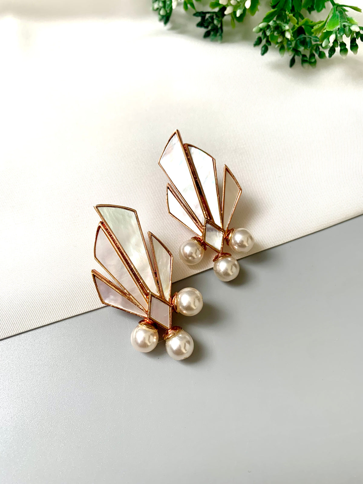 A pair of Art Deco style earrings featuring fan-shaped mother-of-pearl inlays in rose gold-tone metal, accented with small white pearls, displayed on a light gray and white surface with a touch of green foliage in the corner.