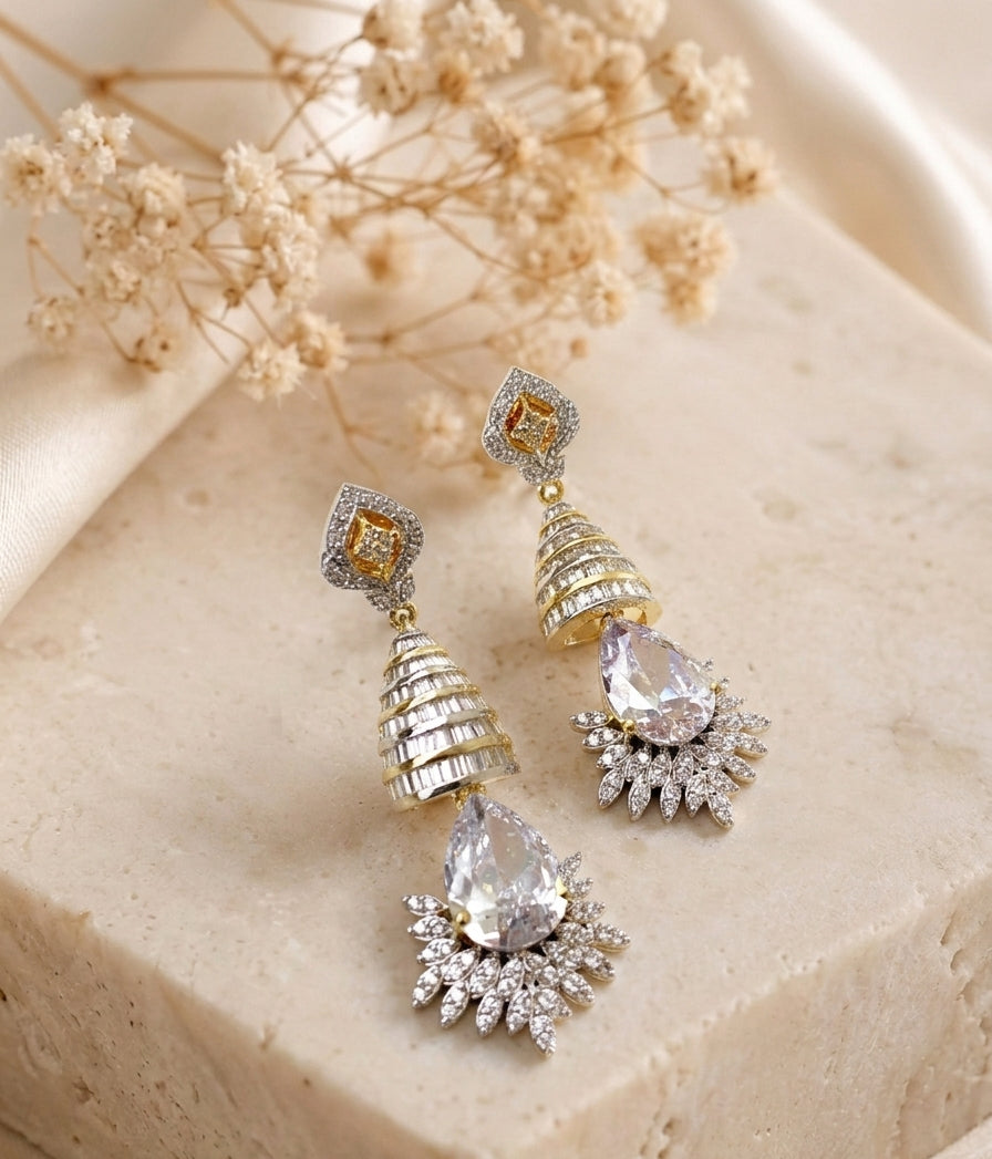 A pair of gold and silver drop earrings with clear gemstones, displayed on a beige stone block with dried flowers in the background.