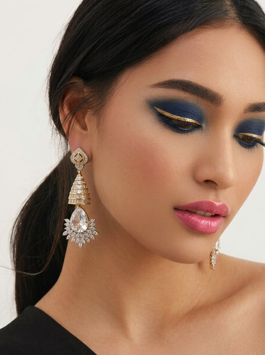 A close-up of a woman wearing a large gold and crystal chandelier drop earring, with matching earrings partially visible on her other side. She has dramatic blue and gold eyeshadow and is wearing a black top against a clean white background.