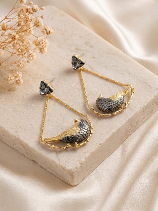 A pair of gold and silver dangling earrings displayed on a beige stone block with cream satin fabric and dried flowers in the background.