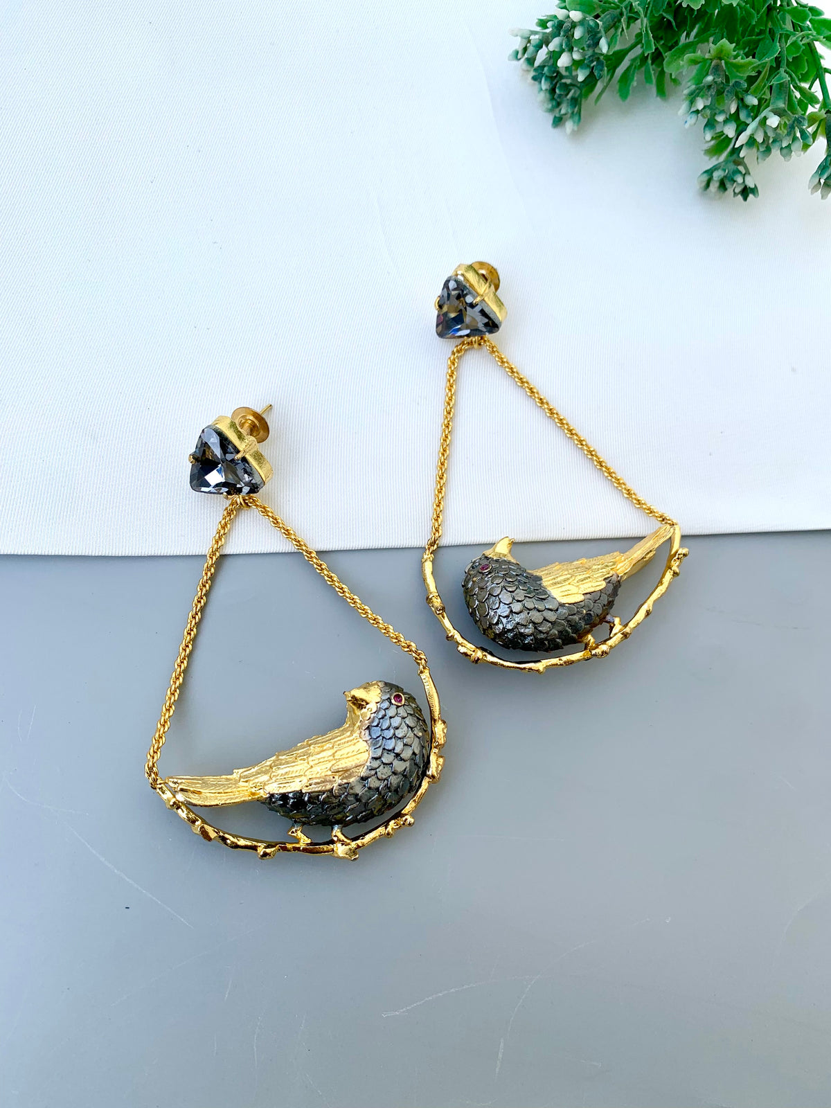 A pair of gold and black bird drop earrings with textured scales, hanging from gold chains with heart-shaped posts, displayed on a split white and gray surface with a small green plant in the corner.