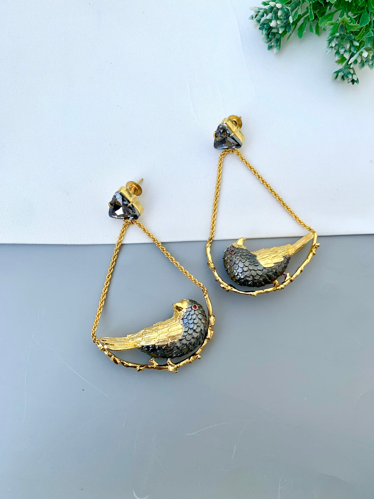 A pair of gold and black bird drop earrings displayed on a split white and gray surface with a small green plant in the corner.