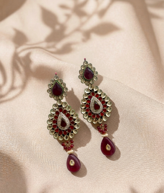 A pair of ornate gold-toned Indian-style dangle earrings with deep red stones and silver pavé detailing, displayed on a beige satin fabric with soft leaf shadows.