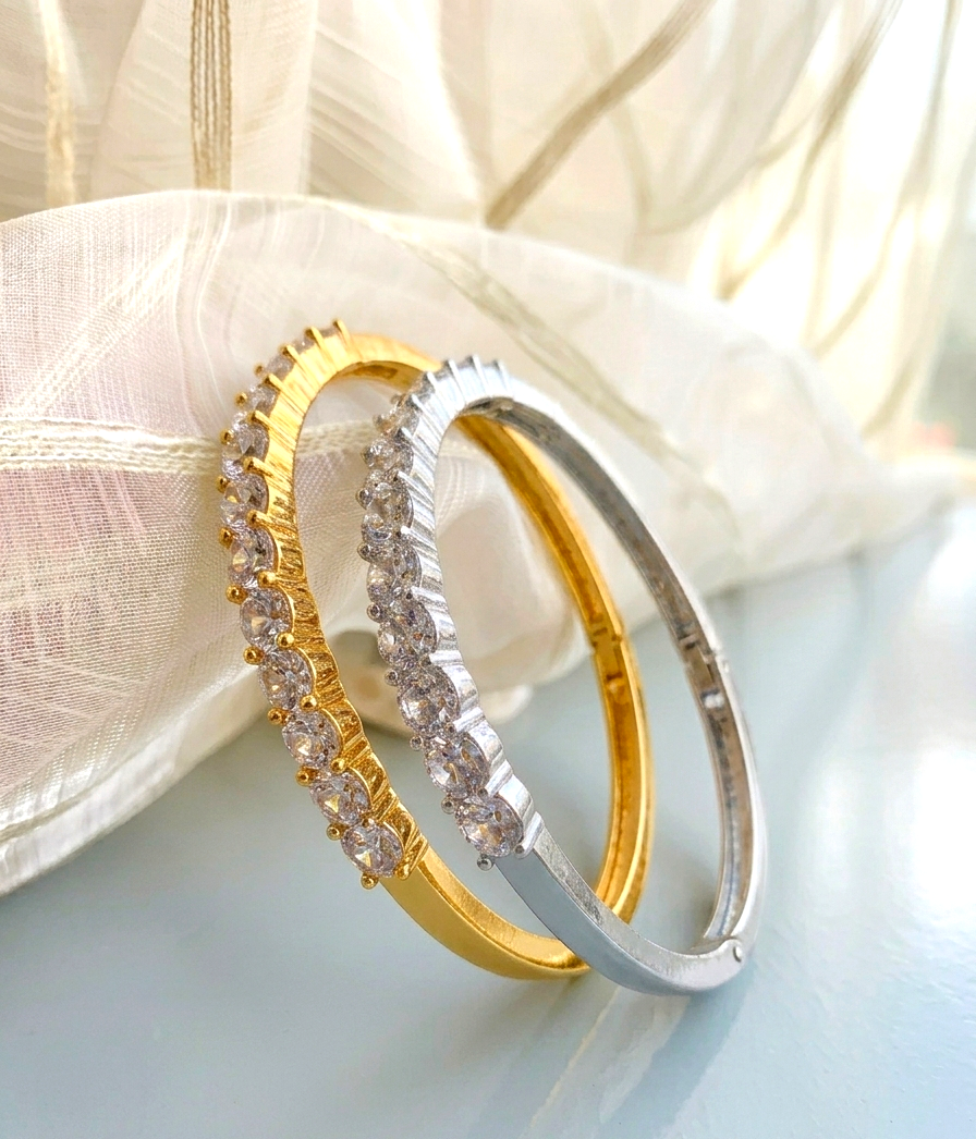 Two slim bangle bracelets, one yellow gold and one silver, both featuring rows of clear pavé-set stones, displayed on a light blue surface with draped white fabric in the background.