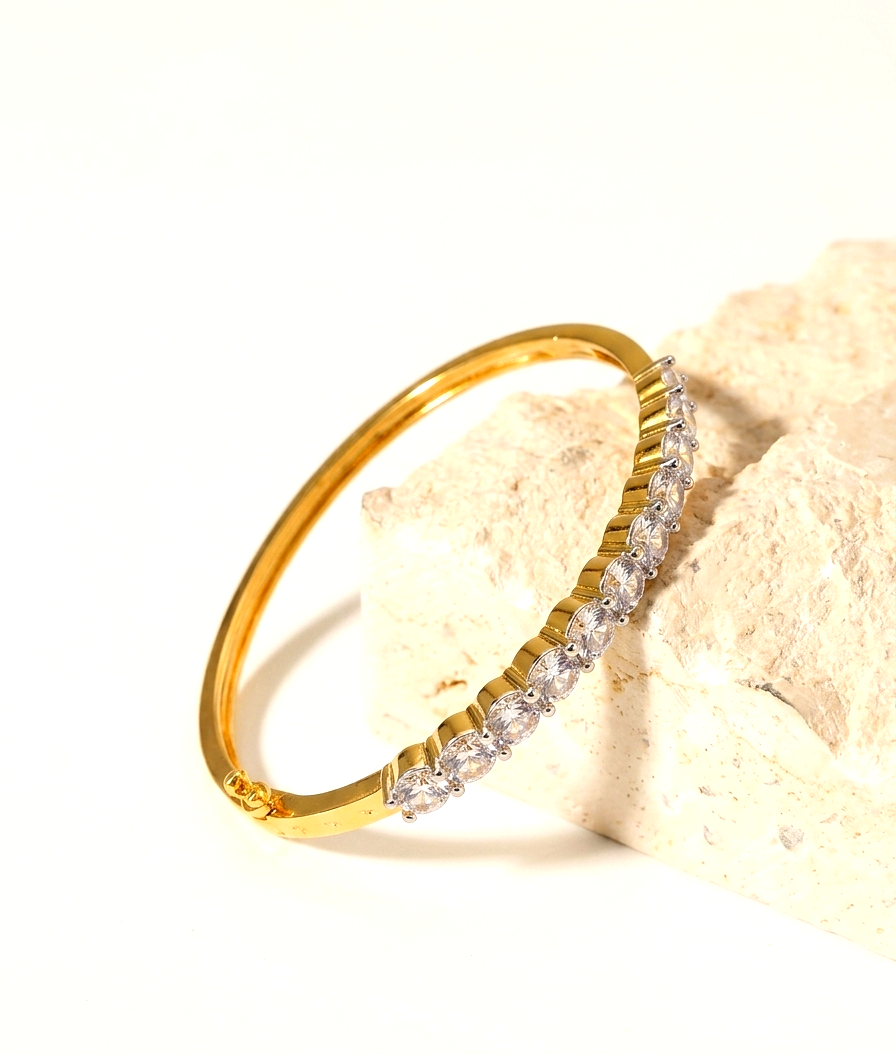 A gold bangle bracelet with a row of clear marquise-cut gemstones, resting on a textured beige stone prop against a bright white background.