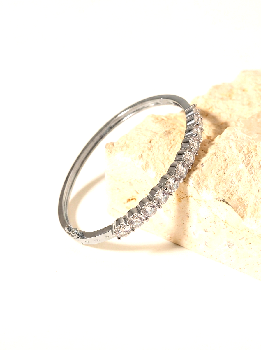 A slim silver-toned bangle bracelet with a row of clear stones along one side, resting on a textured beige stone prop against a bright white background.