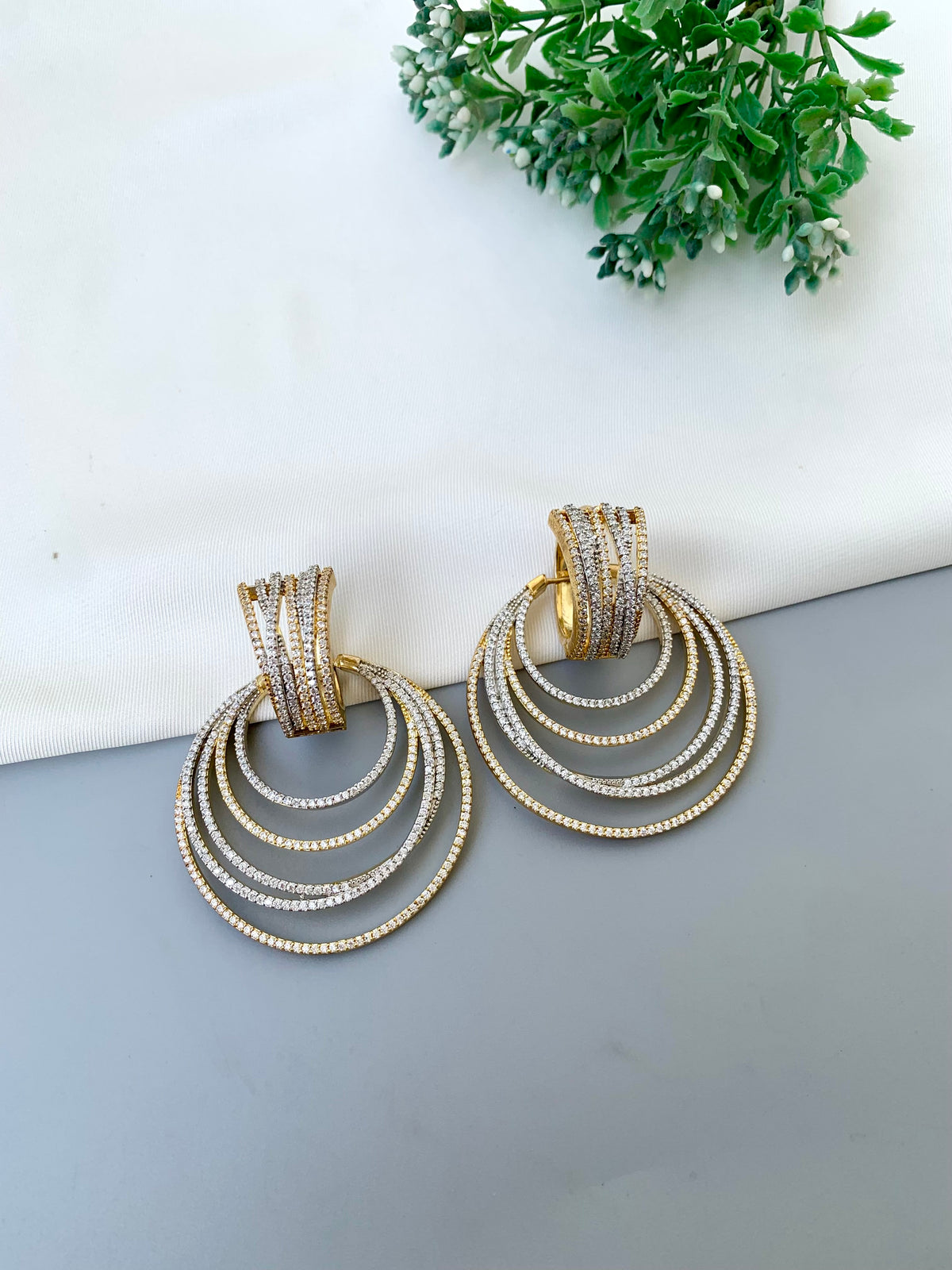 A pair of gold-toned statement earrings with multiple concentric hoop rings, each set with small clear stones, displayed on a gray surface with a white background and green foliage in the corner.