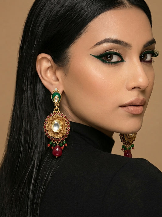 A close-up of a woman with long black hair wearing a large ornate gold-toned dangle earring with green, red, and white details, against a warm beige background.
